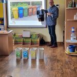 Przed ekranem znajdują się pojemniki do segregacji odpadów_ a obok ekranu stoi mężczyzna z koszem na śmieci w ręku.
