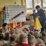 Grupa dzieci siedzi naprzeciwko planszy z pięciolinią i nutkami_ obok dziewczynka wyjęła z worka żółtą nutkę.