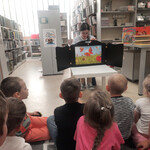 Bibliotekarka pokazuje dzieciom teatrzyk kamishibai_w którym gąsienica z poczwarki zamienia się w motyla.