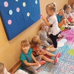 Rząd dzieci siedzące pod tablicą z przyczepionymi kolorowymi kółkami_ dziewczynka stojąca przy tablicy