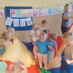 Dzieci stoi na środku chusty, inne okręcają chustę wokół nich, w tle napis dzień przedszkolaka