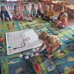 Dzieci siedzą na dywanie_ a dziewczynka kładzie na macie element drogi dla samochodu