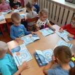 Przedszkolaki siedzą przy stolikach i oglądają książeczki.