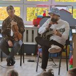 Na krzesłach siedzi dwóch muzyków_ jeden z nich trzyma skrzypce_ drugi gitarę elektroakustyczną