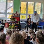 Przed dziećmi stoi pan w białej koszuli lekko pochylony_ obok niego na krzesłach siedzi troje artystów