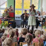 Przed dziećmi stoi pani w kapeluszu na głowie_ trzymająca mikrofon_ a za nią na krzesłach siedzi 3 muzyków