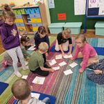 Grupa dzieci znajduje się na dywanie wokół rozrzuconych kartek z brazkami.