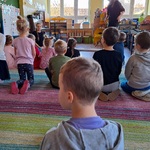 Dzieci siedzą na dywanie naprzeciw nich siedzi kobieta i stoi męzczyzna.