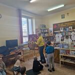 Pani umieszcza emblematy na tablicy_ obok niej stoją 2 chłopcy_ pozostałe dzieci siedzą na dywanie.
