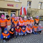 Dzieci w kamizelkach odblaskowych stoją lub kucają przed wejściem do szkoły.