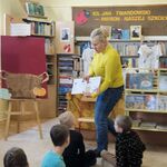 Pani stoi naprzeciwko dzieci_ prezentuje książkę oraz kartę biblioteczną.