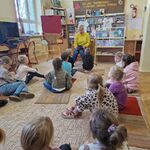 Dzieci siedzą na dywanie_ a naprzeciwko nich siedzi pani na krześle i trzyma książkę.