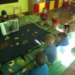 dzieci siedzą wokół dywanu na którym leżą liście drzew_ dziewczynka pokazuje książkę.
