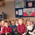Grupa dzieci siedzi na dywanie_ za nimi na ścianie wisi mapa Polski_ napis Polska i Godło Polski.