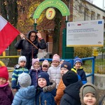 Grupa dzieci stoi przed bramką do przedszkola_ z tyłu stoi pani_ trzyma flagę.