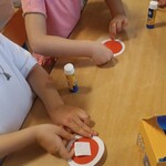 Dzieci siedzą przy stole_ przyklejają biało-czerwone elementy tworząc kotyliony.