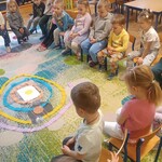 grupa dzieci siedzi na krzesłach_ pani rozkłada na dywanie obręcze z bibuły.