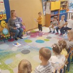 grupa przedszkolaków na krzesłach_ pod ścianą siedzi pani z książką.