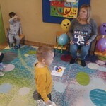 pani trzyma książkę z napisem kosmos_ obok niej na krzesłach siedzą kosmici wykonani z balonów_ obok dzieci na krzesełkach.