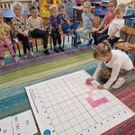 Dzieci siedzą na krzesełkach_ a dziewczynka na macie układa obrazek z różowych kartoników.