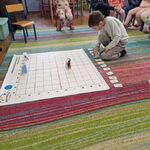 Chłopiec jest na środku dywanu przed matą do kodowania_ trzyma w ręku Genibota i dotyka kart do programowania.