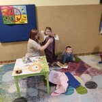 pani owija uśmiechniętą dziewczynkę materiałem_ obok siedzi chłopiec.