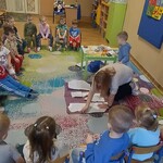 grupa dzieci siedzi na krzesełkach_ pani na dywanie rozkłada wycięty papier na materiale.