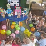 Dzieci siedzą na dywanie_ przed nimi leżą kolorowe balony_ a na stole stoją roboty.
