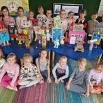Na stołach znajdują się roboty_ grupa dzieci stoi za stołem_ a kilkoro dzieci siedzi przed stołem.