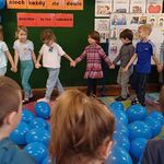 Na środku dywanu leżą balony_ dzieci trzymając się za ręce idą dookoła balonów.