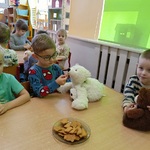 Przedszkolaki siedzą przystolikach i częstują misie miodowymi ciasteczkami.