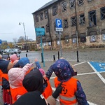 Dzieci stoją na chodniku_ przed nimi_ po drugiej stronie ulicy ceglany budynek osłonięty siatką z oknami zabitymi deskami.
