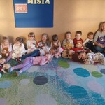 grupa dzieci siedzi pod ścianą_ każde trzyma misia_ w tle napis na tablicy dzień misia.