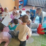 na dywanie grupa dzieci z panią_ trzymają kartki.