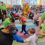 Troje dzieci w bokserskich rękawicach_ przed nimi trzech zawodników w pozycji ochronnej_ trenerka przechodzi za dziećmi.