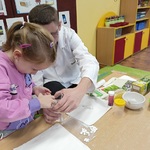 Dziewczynka z pomocą farmaceuty nabiera wody do strzykawki.