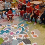 dzieci siedzą na krzesłach_ na dywanie leżą kartki z namalowanymi prezentami_ jedno dziecko bierze kartkę z dywanu.