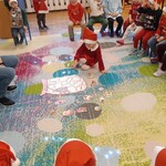 dzieci siedzą na krzesłach_ po lewej pani_ na dywanie dziecko dopasowuje obrazek do kształtu na worku.