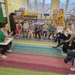 Kobieta siedzi na krzesełku naprzeciwko dzieci_ trzyma na kolanach otwartą książkę.
