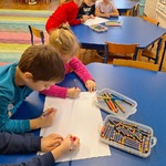 po troje dzieci siedzi przy stolikach rysując na wspólnych kartkach.