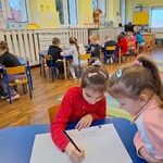 dwie dziewczynki siedząc przy stoliku rysują_ w tle widać pozostałe grupki dzieci przy stolikach i włączony ekran.