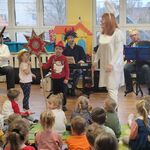 Dwoje dzieci trzyma gwiazdę_ kobieta w stroju anioła stoi obok dzieci_ w tle muzycy grają na instrumentach.