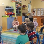 Pani trzyma książkę_ przewraca stronę_ dzieci siedzą w rzędach_ słuchają opowiadania