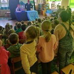 Dzieci siedzą na krzesełkach_ oglądają teatrzyk_ trójka z nich ogląda na stojąco_ zwrócone są przodem do sceny.