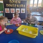 Kilkoro dzieci siedzi przy stoliku i obserwuje przez lupę dinozaury ukryte w kostkach lodu.