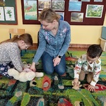 Dziewczynka i chłopiec pod instrukcją kobiety ćwiczą na na misiach umiejętność udzielania pierwszej pomocy.