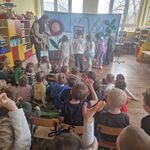 Kilkoro dzieci stoi naprzeciwko aktora i spogląda na rozrzucone na podłodze patyczki.
