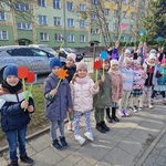 Dzieci trzymając papierowe kwiaty stoją w szeregu na tle bloku.