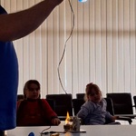 dwie dziewczynki siedzą na krzesłach_ pracownik PGE trzyma żarówkę zasilaną przez małą elektrownię stojąca na stole.