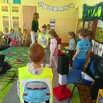 Przedszkolaki w kamizelkach imitujących pojazdy i piesi uczestniczą w ruchu drogowym na makiecie.